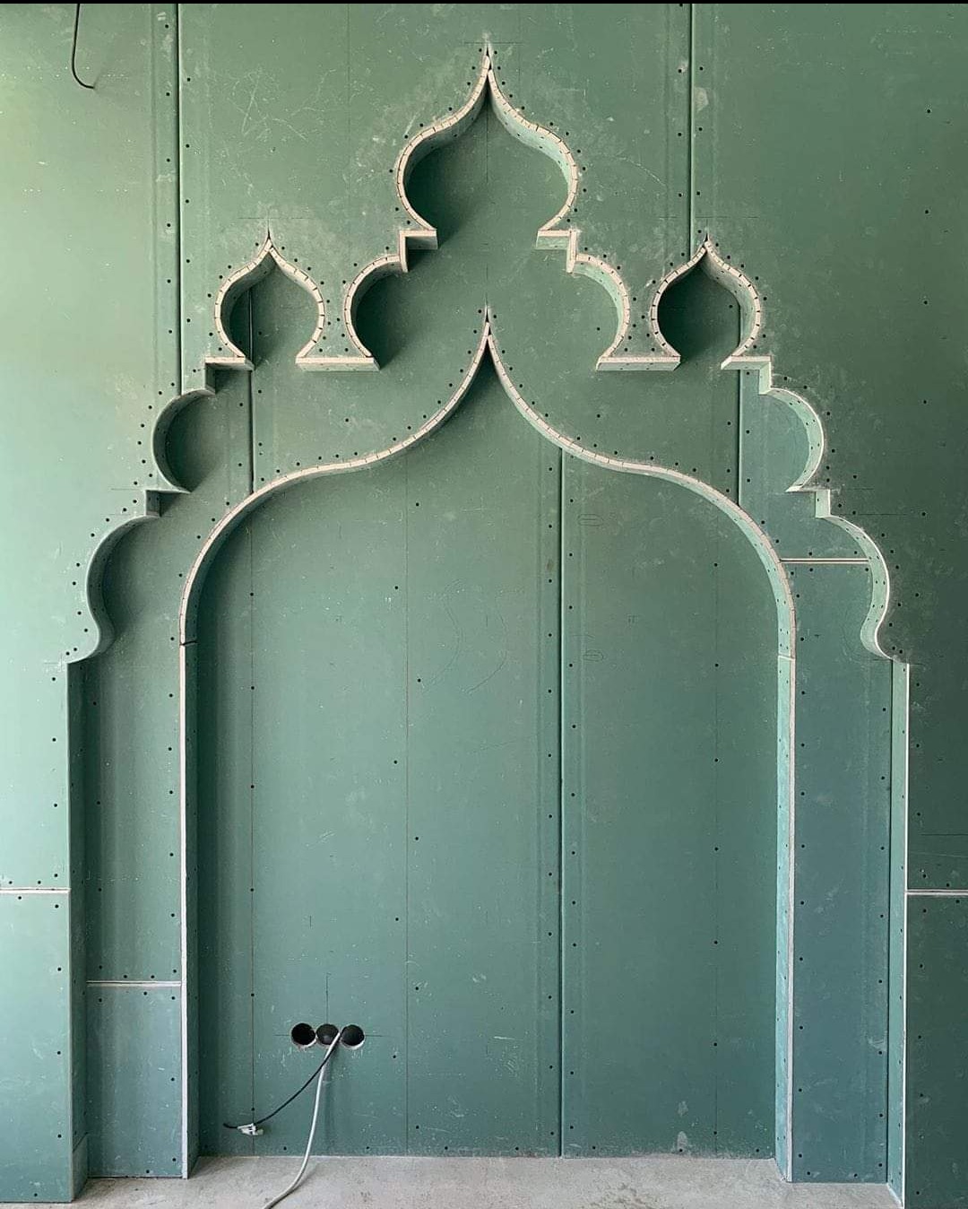 Nietypowa konstrukcja z płyty karton-gips w kolorze zielonym, tworząca dekoracyjny łuk z orientalnymi motywami. Widoczne otwory na gniazdka elektryczne i przewody.