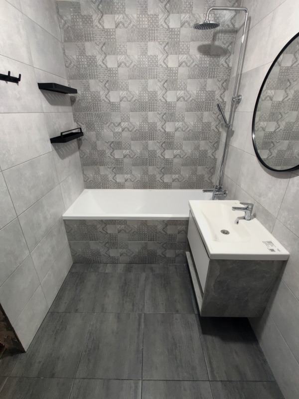 Wykończona łazienka w szarościach: płytki patchworkowe nad wanną, szara podłoga, biała umywalka z szafką i czarne półki ścienne. Nowoczesny design.