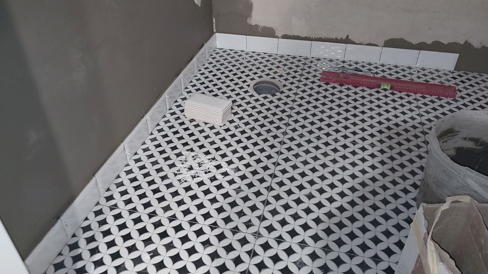 Układanie wzorzystej, czarno-białej glazury podłogowej w łazience. Widoczny poziomica, zapas płytek i wiadro z zaprawą. Ściany przygotowane do dalszych prac.