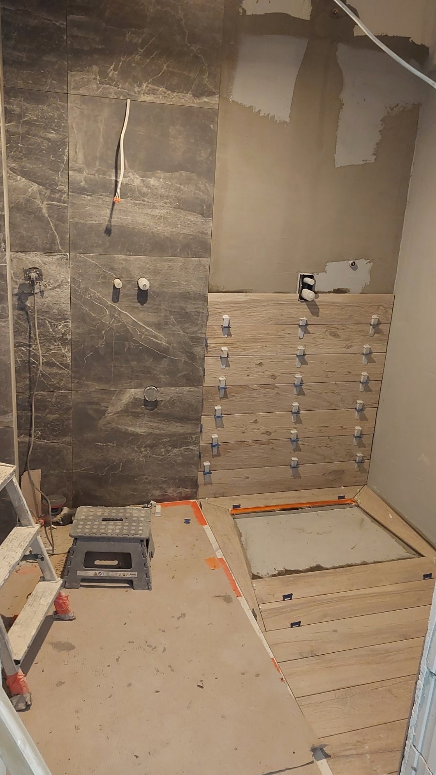 Układanie płytek imitujących drewno w łazience, widoczne krzyżyki dystansowe. Część ściany wykończona płytkami w odcieniach szarości. W tle drabina i surowa ściana.