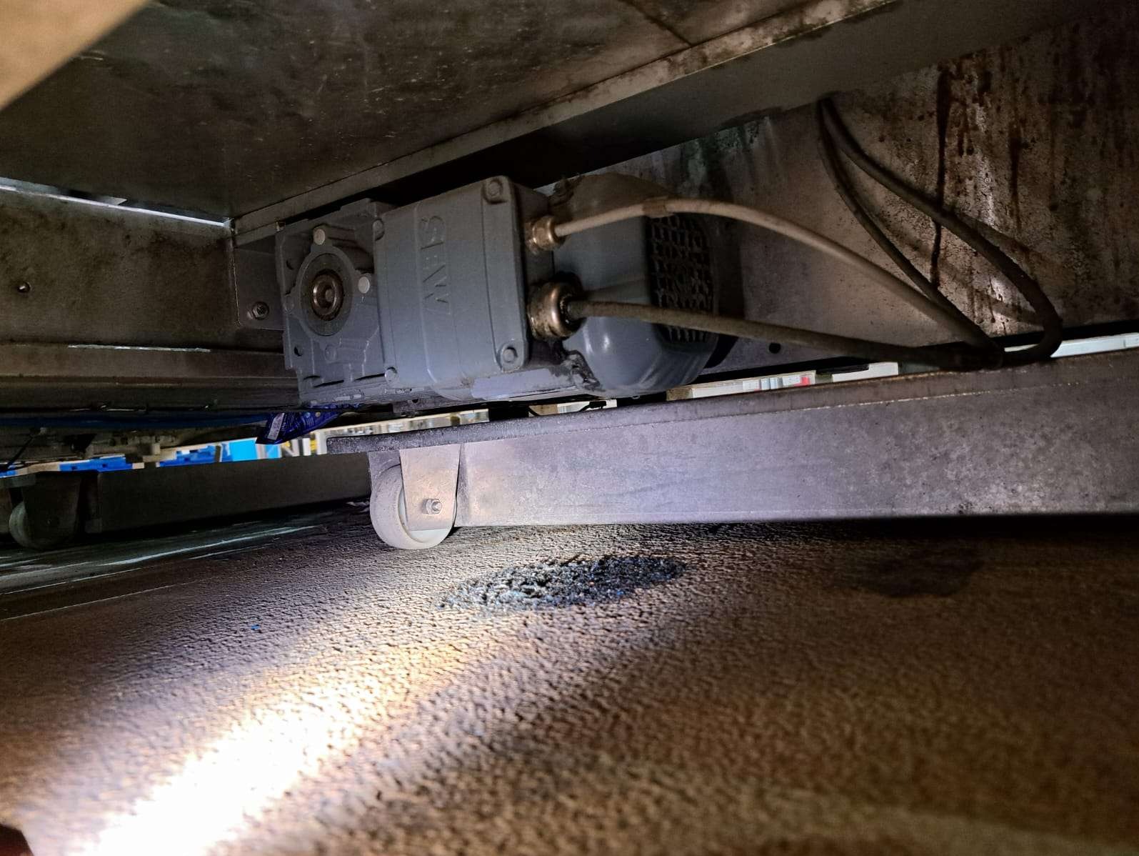 Szary silnik elektryczny SEW zamontowany pod metalową konstrukcją na kółkach, oświetlony od dołu. Widoczne przewody i zabrudzenia na podłożu.