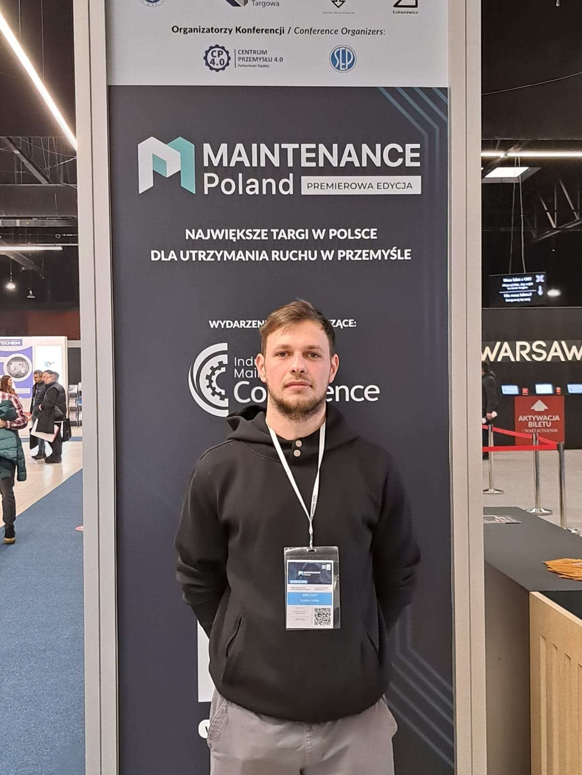 Mężczyzna na tle stoiska Maintenance Poland, targi dla utrzymania ruchu w przemyśle. Ubrany w bluzę z identyfikatorem. Widoczne logo i hasło targów.