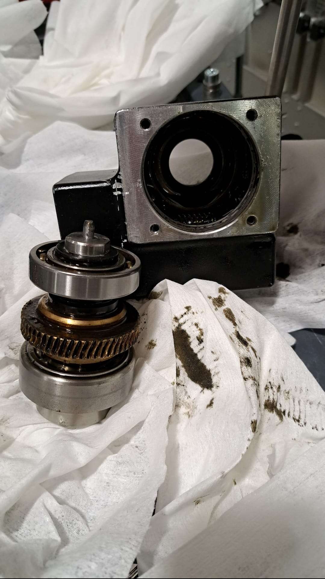 Rozłożony mechanizm przekładni zębatej i łożysk na białej, zabrudzonej olejem ściereczce. Widoczny element obudowy. Precyzyjny demontaż maszyny.