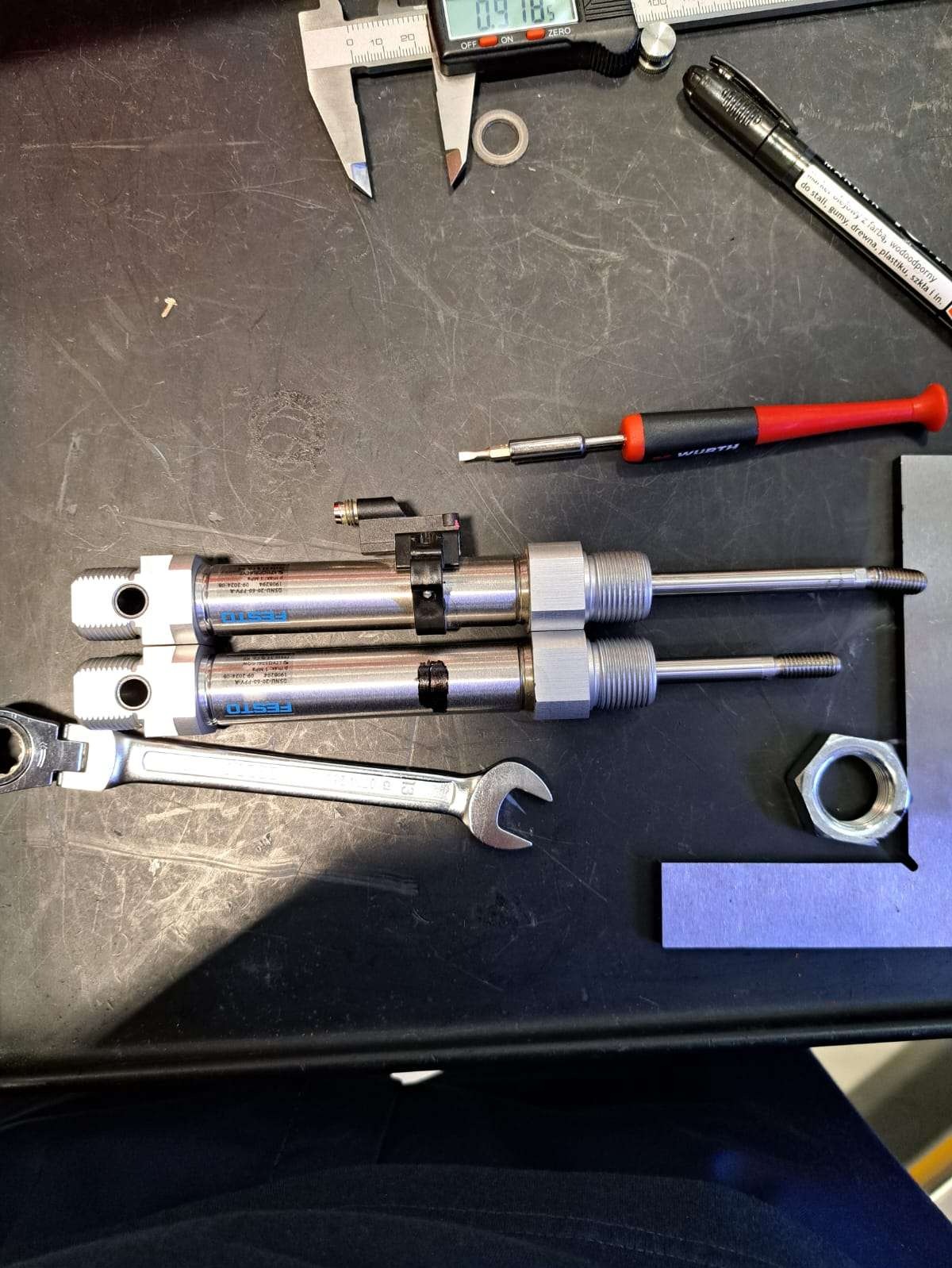 Dwa siłowniki pneumatyczne Festo leżą obok klucza, suwmiarki i śrubokręta Wurth na czarnym stole warsztatowym. Widoczny fragment kątownika i nakrętka.