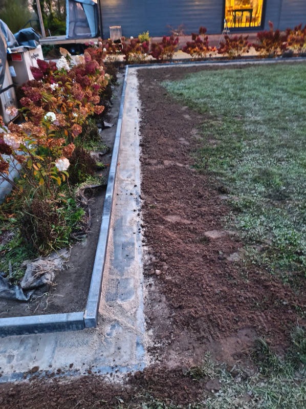 Krawężnik betonowy oddzielający rabatę z hortensjami od trawnika z przygotowaną ziemią. Widoczne ślady prac ziemnych i elementy konstrukcyjne.