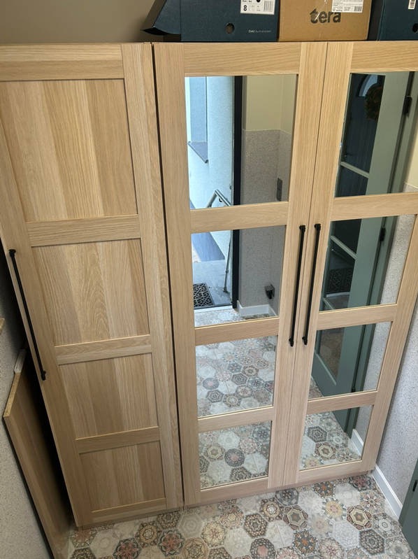 Jasna, drewniana szafa z lustrami w drzwiach, odbijająca wzorzystą podłogę. Widoczne czarne uchwyty i kartonowe pudła na górze. Ujęcie z bliska, wnętrze mieszkania.