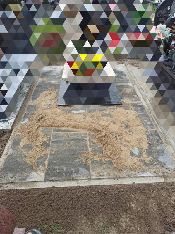 Wykończony nagrobek z szarej kostki, częściowo zasypany piaskiem. Widoczne są sąsiednie nagrobki i zieleń w tle. Ujęcie z góry, z bliska.