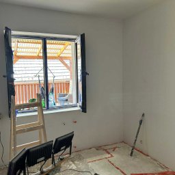 Usługi Remontowo-Wykończeniowe Dawid Wędrzyk - Wnętrze pokoju po szpachlowaniu ścian, widoczne narzędzia, drabina i okno z widokiem na taras. Ściany przygotowane do dalszych prac wykończeniowych.