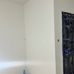 Usługi Remontowo-Wykończeniowe Dawid Wędrzyk - Białe, świeżo pomalowane ściany w rogu pokoju, zabezpieczone drzwi folią malarską. Na podłodze widoczne ślady farby i butelka wody mineralnej.