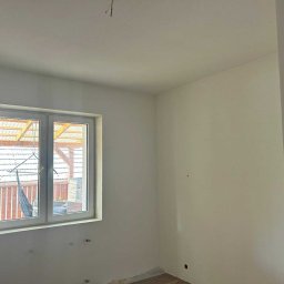 Usługi Remontowo-Wykończeniowe Dawid Wędrzyk - Jasne wnętrze po malowaniu ścian na biało, widoczne gniazdka elektryczne i okno z widokiem na zadaszony taras. Minimalistyczny styl wykończenia.