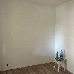 Usługi Remontowo-Wykończeniowe Dawid Wędrzyk - Pomalowane na biało ściany w rogu pokoju z drewnianą podłogą. Na taborecie butelka wody, szpachelka i puszka z farbą. Widoczne gniazdko elektryczne.