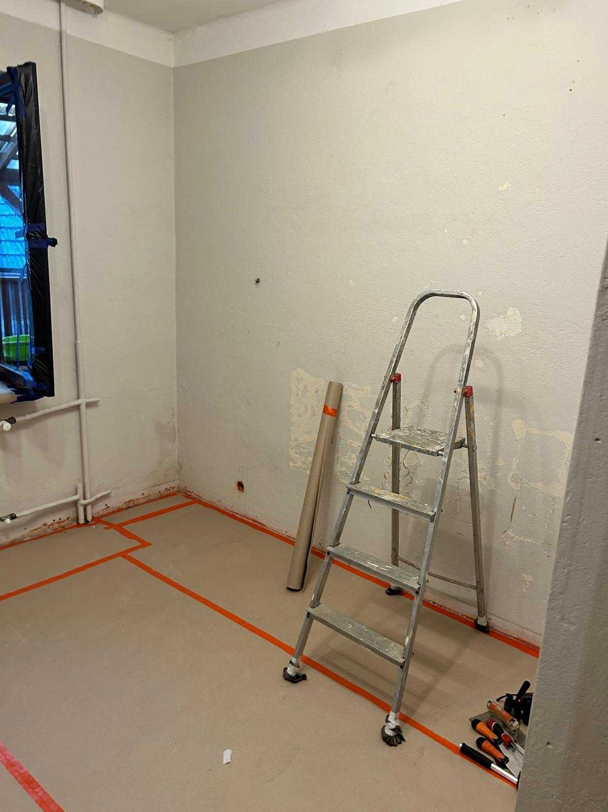 Pokój w trakcie przygotowań do malowania: ściany z widocznymi śladami po szpachlowaniu, zabezpieczona podłoga taśmą malarską, drabina i narzędzia malarskie.