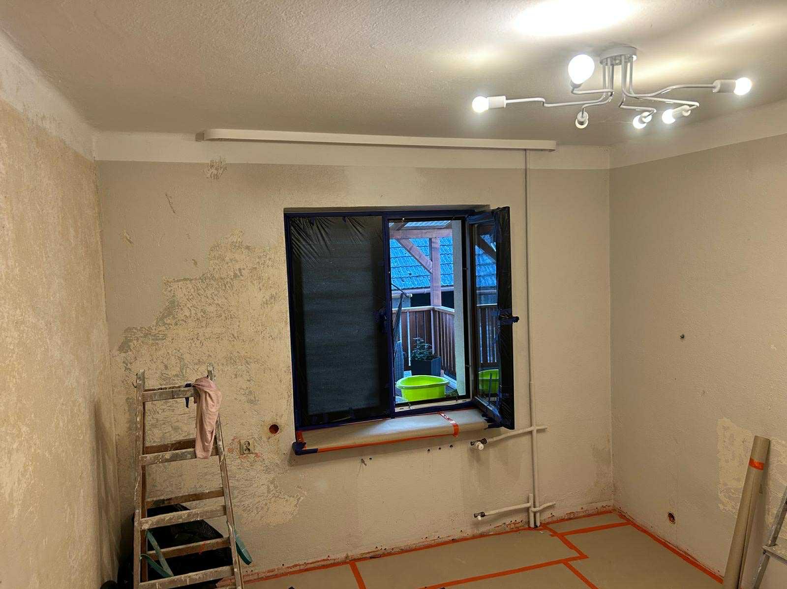 Pokój w trakcie przygotowań do malowania: ściany z resztkami starej farby, okno zabezpieczone folią, drabina i taśmy malarskie na podłodze.
