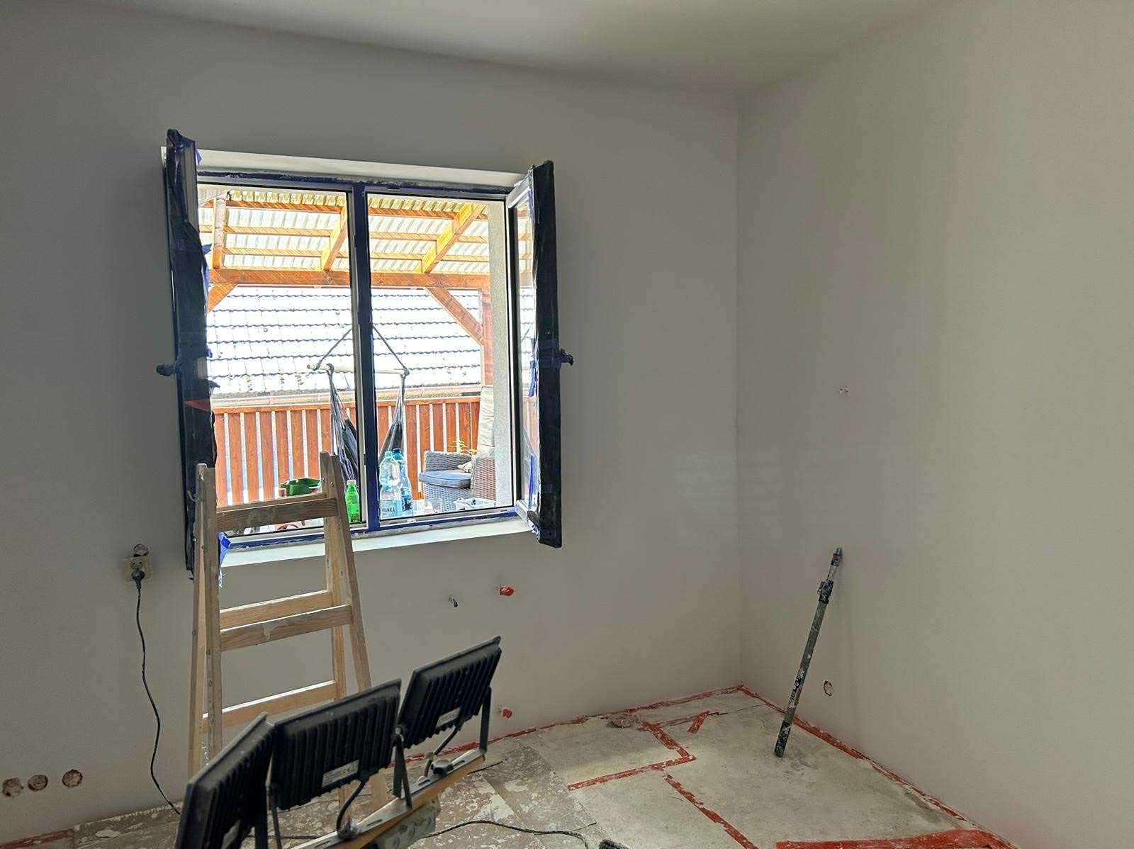 Wnętrze pokoju po szpachlowaniu ścian, widoczne narzędzia, drabina i okno z widokiem na taras. Ściany przygotowane do dalszych prac wykończeniowych.