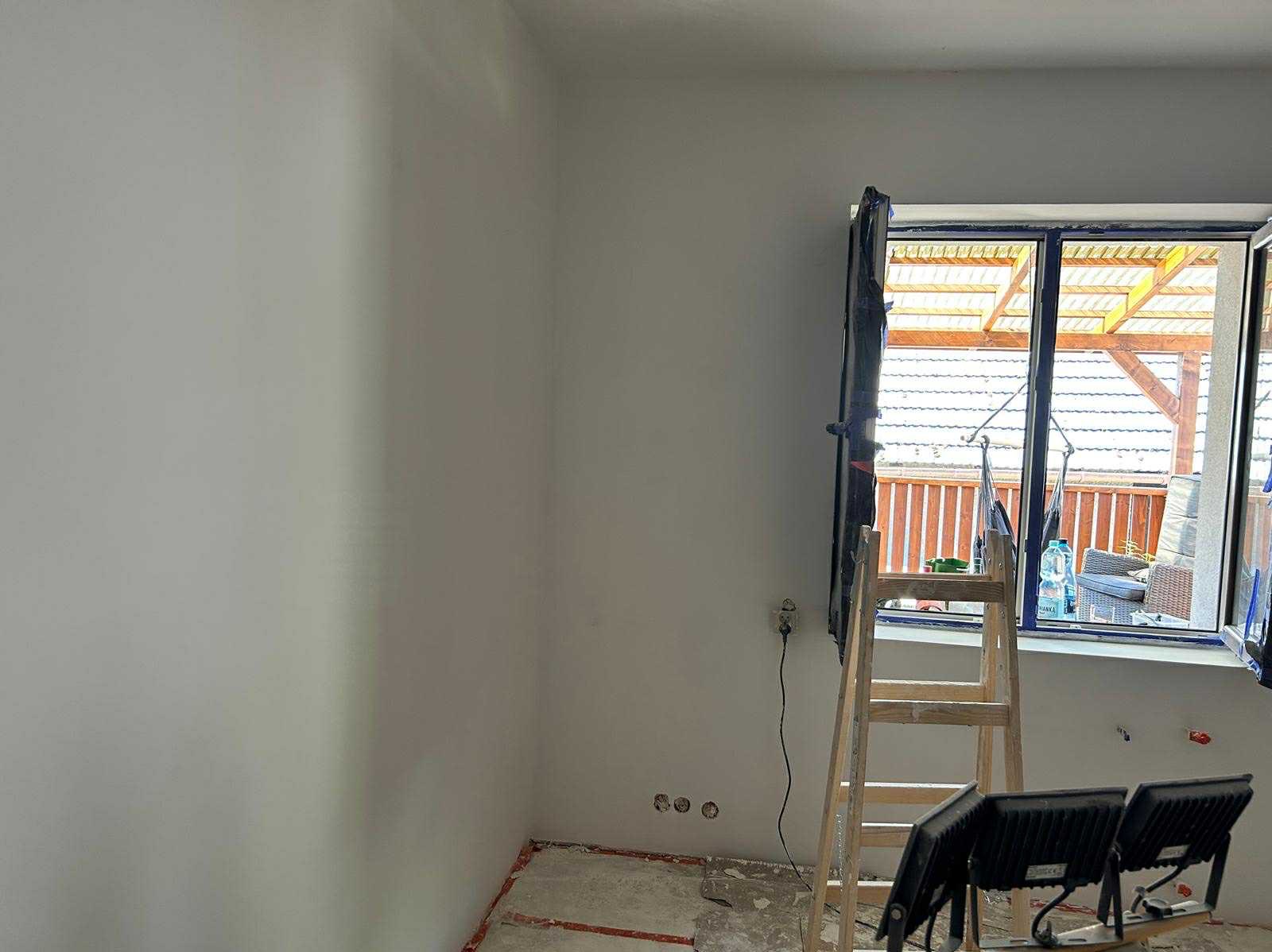 Wnętrze pokoju po szpachlowaniu ścian, przygotowane do malowania. Widoczne ślady po gniazdkach, drabina i lampy halogenowe. Okno z widokiem na balkon.