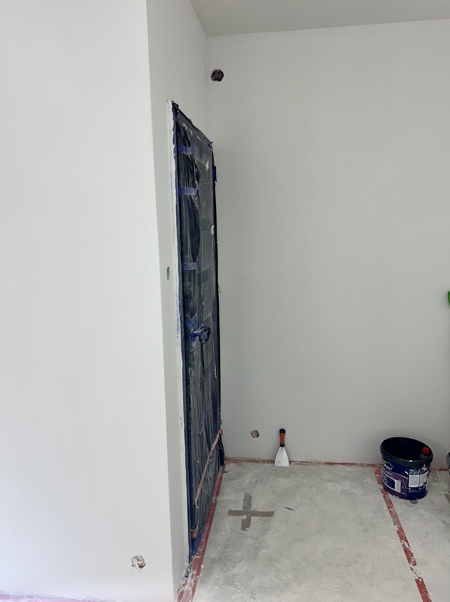 Białe, świeżo pomalowane wnętrze z zabezpieczonymi drzwiami folią, widoczne narzędzie i wiadro z farbą. Minimalistyczny kadr, surowy beton na podłodze.
