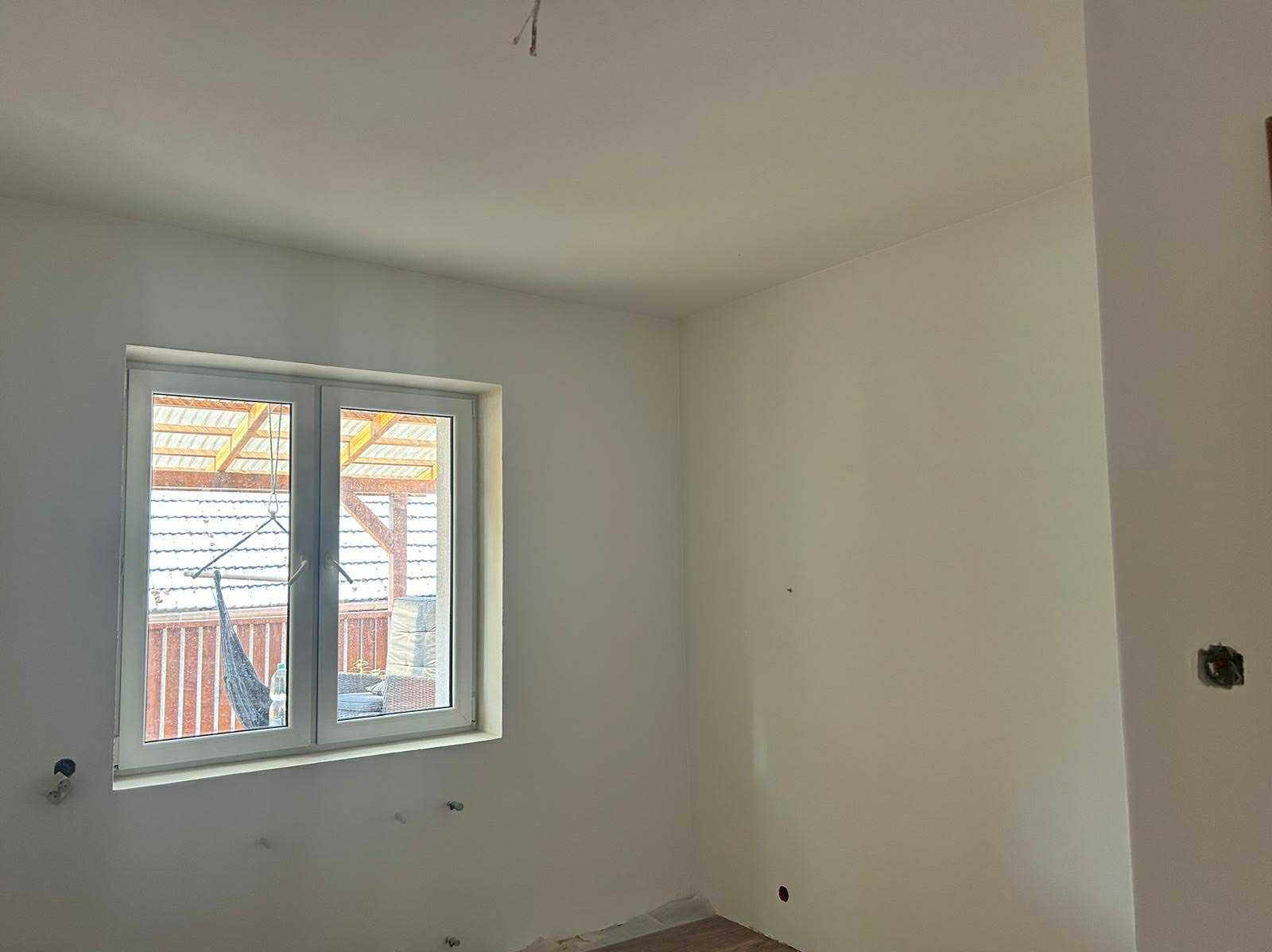 Jasne wnętrze po malowaniu ścian na biało, widoczne gniazdka elektryczne i okno z widokiem na zadaszony taras. Minimalistyczny styl wykończenia.