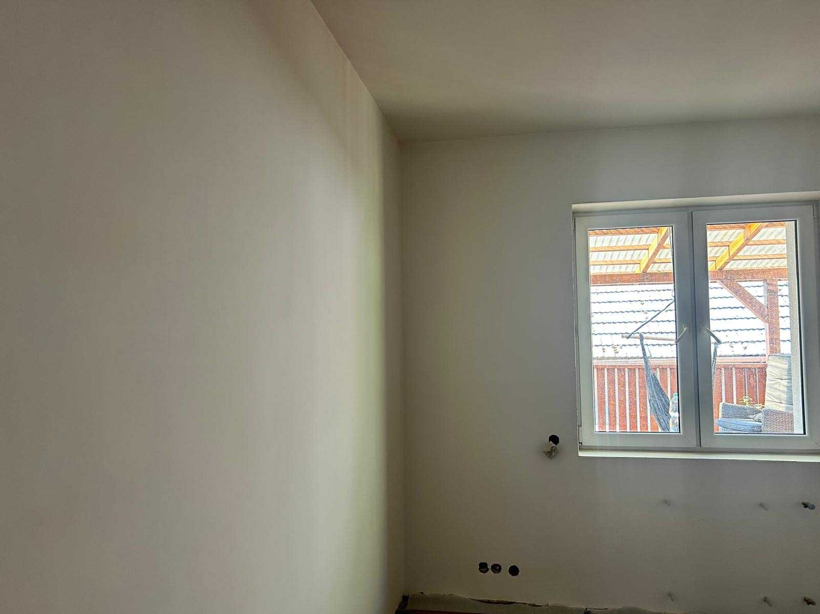 Wnętrze po szpachlowaniu i malowaniu na jasny odcień. Widoczne gładkie ściany, okno z widokiem na zewnątrz i przygotowane punkty elektryczne.
