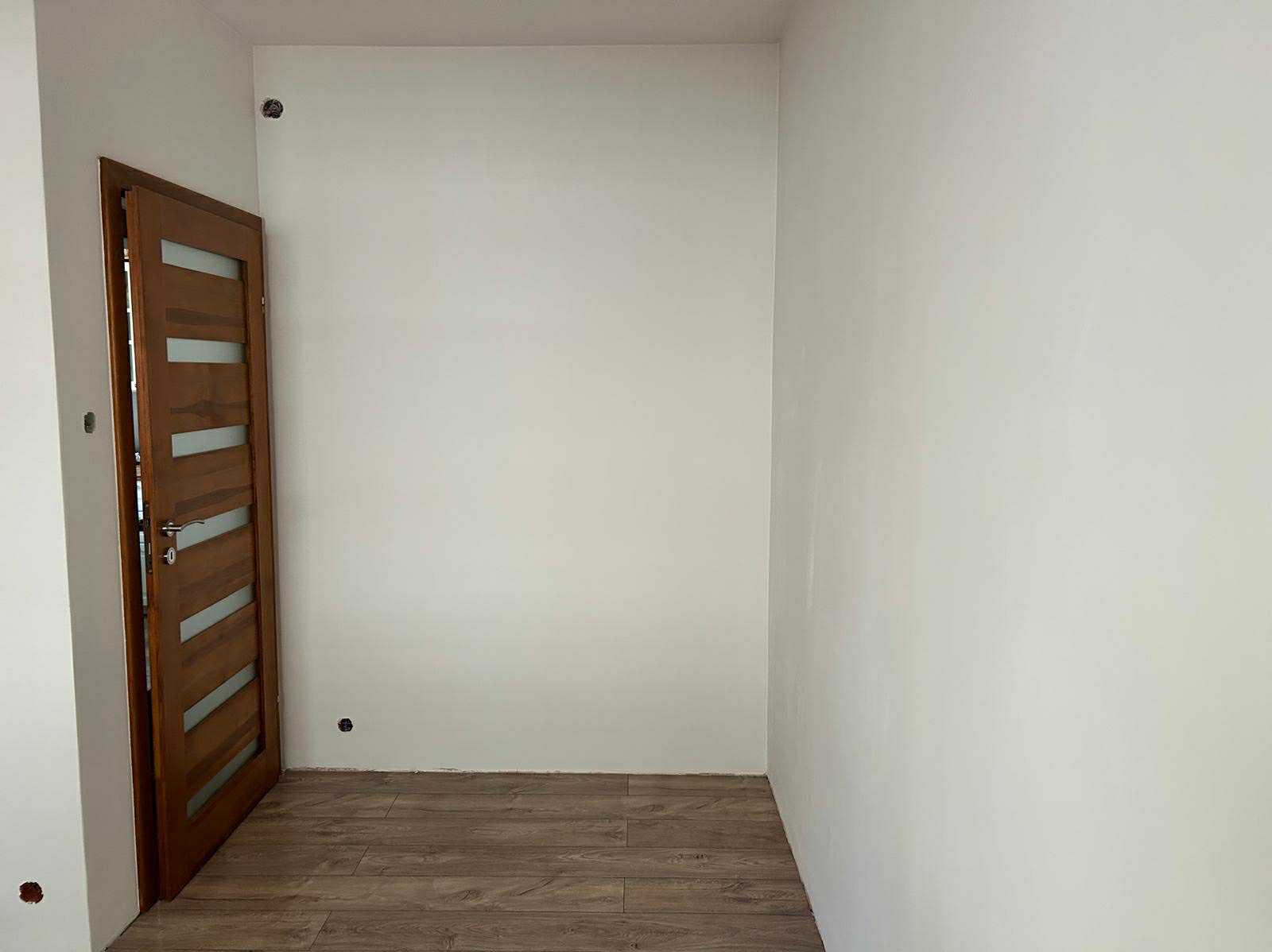 Jasne wnętrze po remoncie: drewnopodobne panele, białe ściany z widocznymi gniazdkami i częściowo otwarte drzwi z przeszkleniami. Minimalistyczny design.