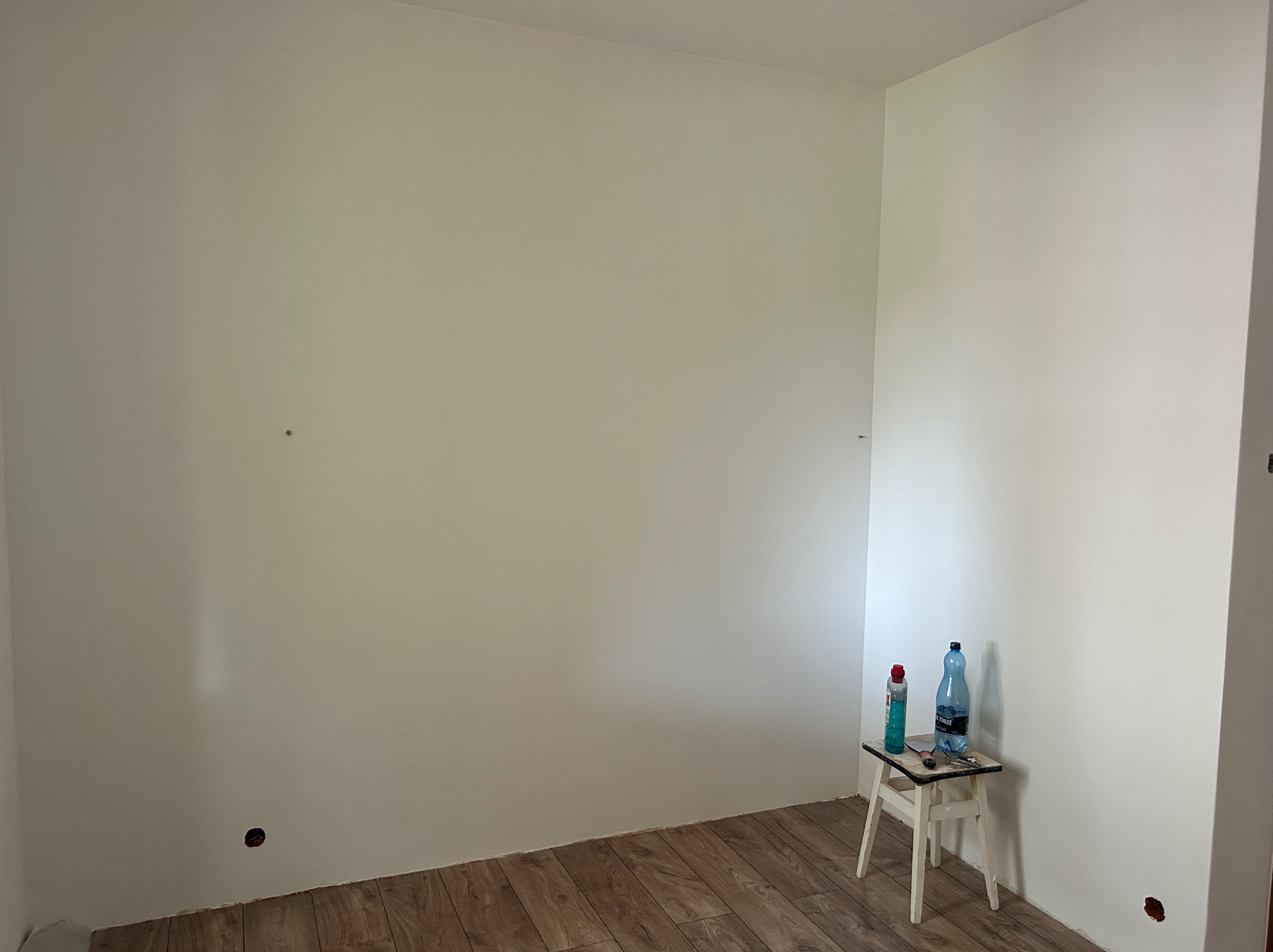 Pomalowane na biało ściany w rogu pokoju z drewnianą podłogą. Na taborecie butelka wody, szpachelka i puszka z farbą. Widoczne gniazdko elektryczne.