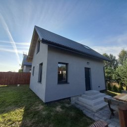 Lilmil Anna Łosińska - Nowoczesny, parterowy dom z szarą elewacją i ciemnym dachem, otoczony zielonym trawnikiem i drewnianym ogrodzeniem. Widoczne schody wejściowe i okno z odbiciem wnętrza.