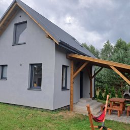 Lilmil Anna Łosińska - Nowoczesny, parterowy dom z szarą elewacją i drewnianą pergolą nad wejściem. W tle zielony ogród i pochmurne niebo. Widoczny hamak i meble ogrodowe.