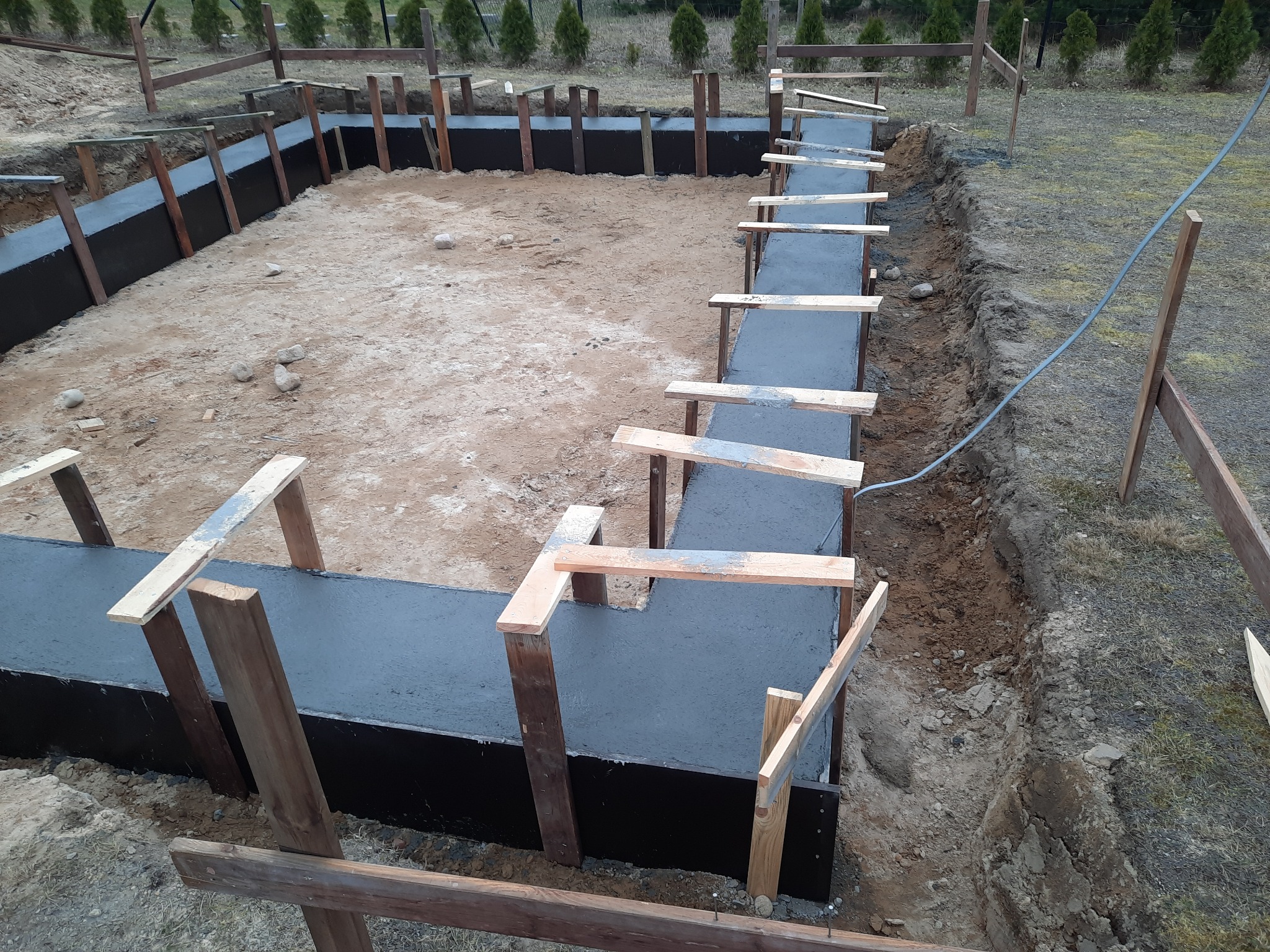 Zalane betonem fundamenty domu jednorodzinnego, zabezpieczone deskowaniem i drewnianymi stemplami. Widok z góry na wykop z widoczną ziemią i roślinnością w tle.