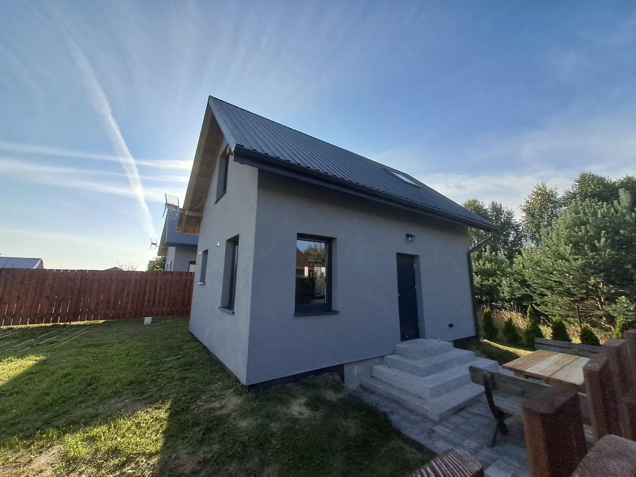 Nowoczesny, parterowy dom z szarą elewacją i ciemnym dachem, otoczony zielonym trawnikiem i drewnianym ogrodzeniem. Widoczne schody wejściowe i okno z odbiciem wnętrza.