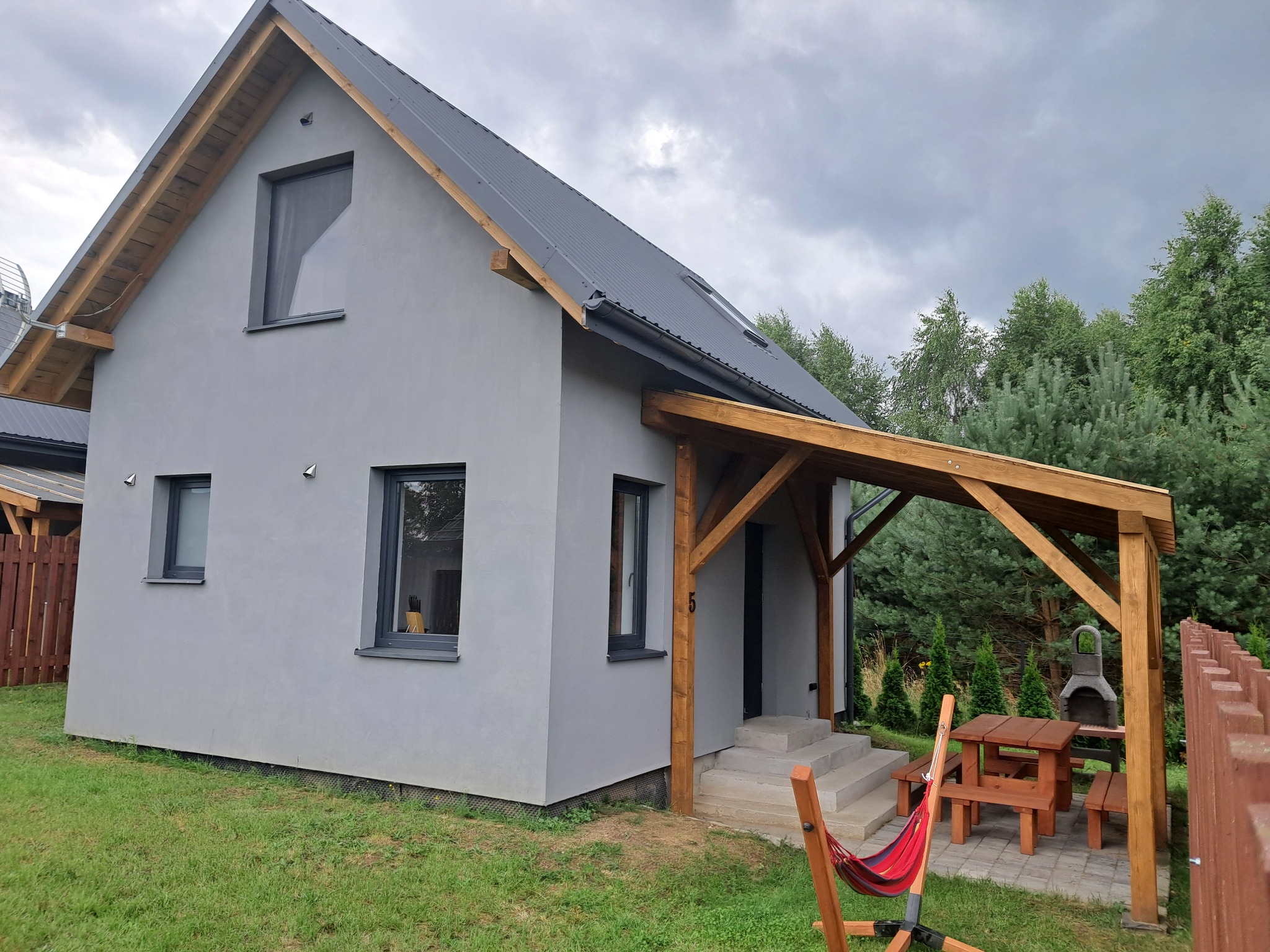 Nowoczesny, parterowy dom z szarą elewacją i drewnianą pergolą nad wejściem. W tle zielony ogród i pochmurne niebo. Widoczny hamak i meble ogrodowe.