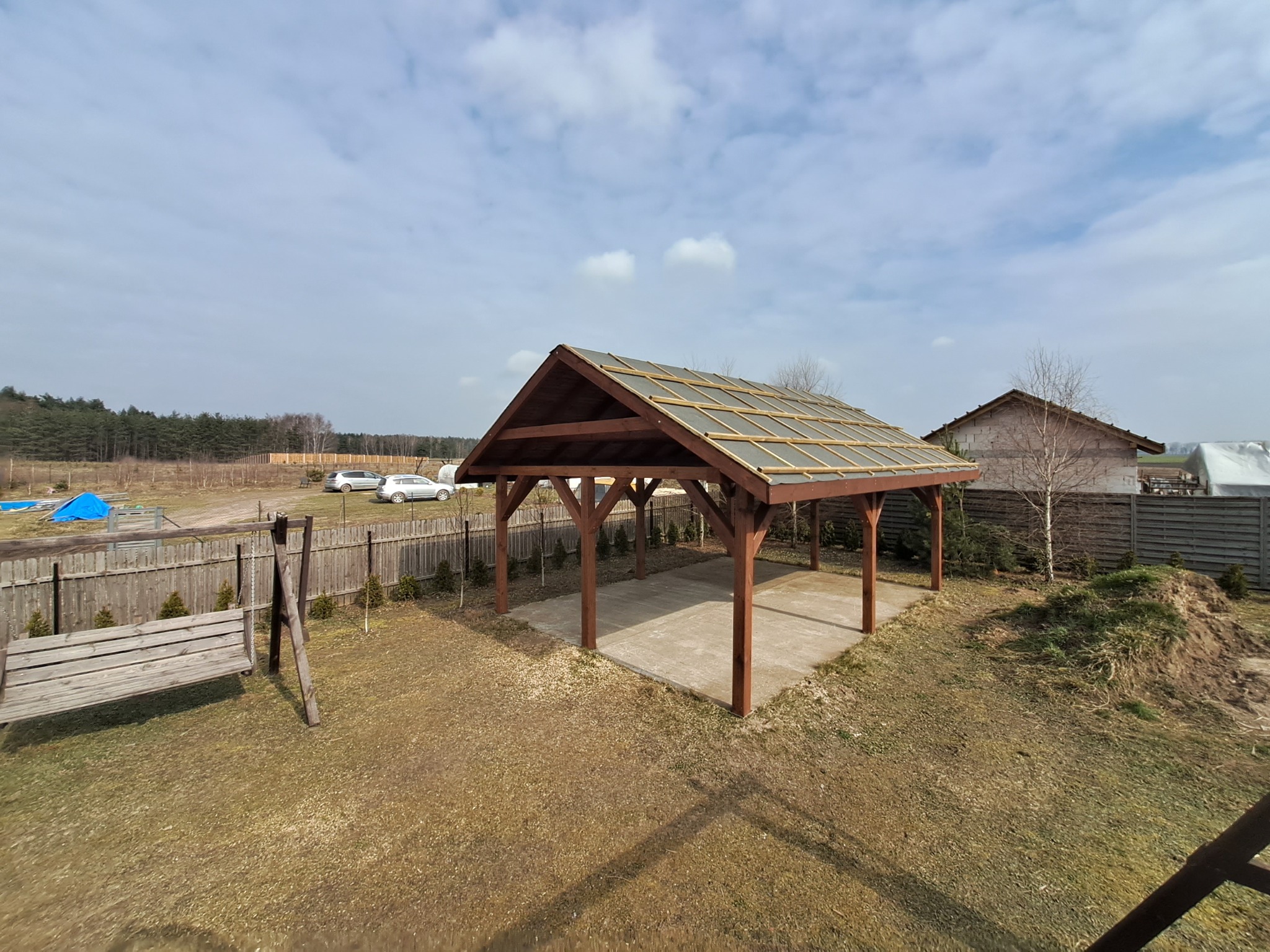 Drewniana altana z betonową podłogą w ogrodzie, widok z góry. W tle dom, drzewa i błękitne niebo z chmurami. Huśtawka ogrodowa po lewej stronie.