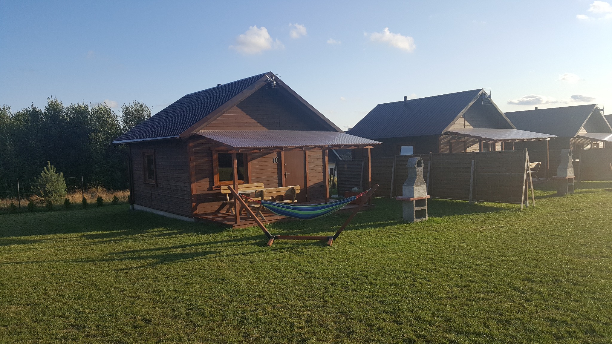 Kompleks drewnianych domków letniskowych z zadaszonymi tarasami, hamakiem i murowanymi grillami na zadbanym trawniku w słoneczny dzień.