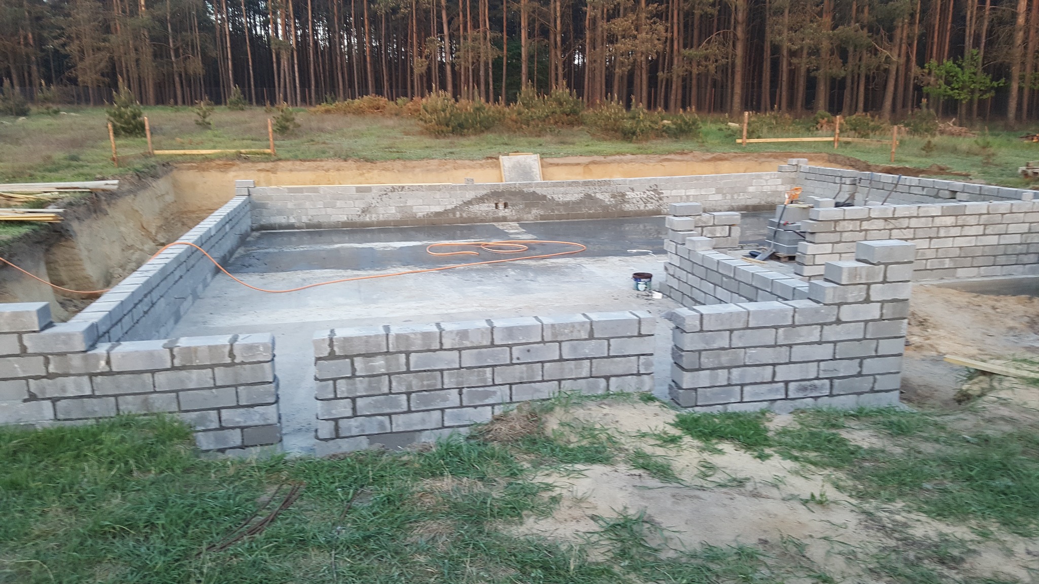 Wznoszenie ścian fundamentowych z bloczków betonowych na wylewce, widoczne narzędzia i pomarańczowy przewód. W tle las i wykopany teren pod budowę.
