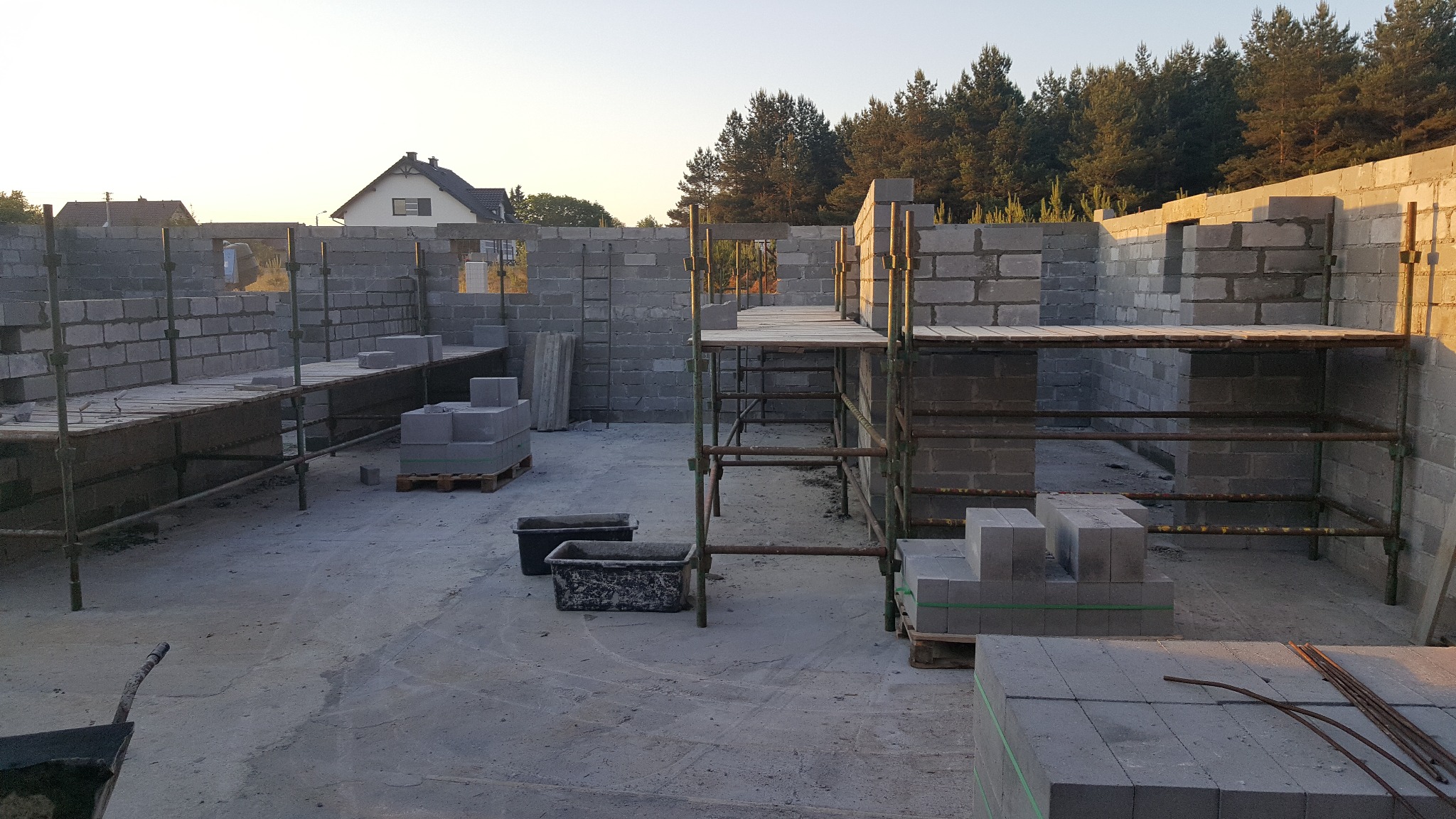 Wznoszenie ścian z bloczków betonowych na budowie domu jednorodzinnego. Widoczne rusztowania i palety z materiałem. W tle dom i drzewa.