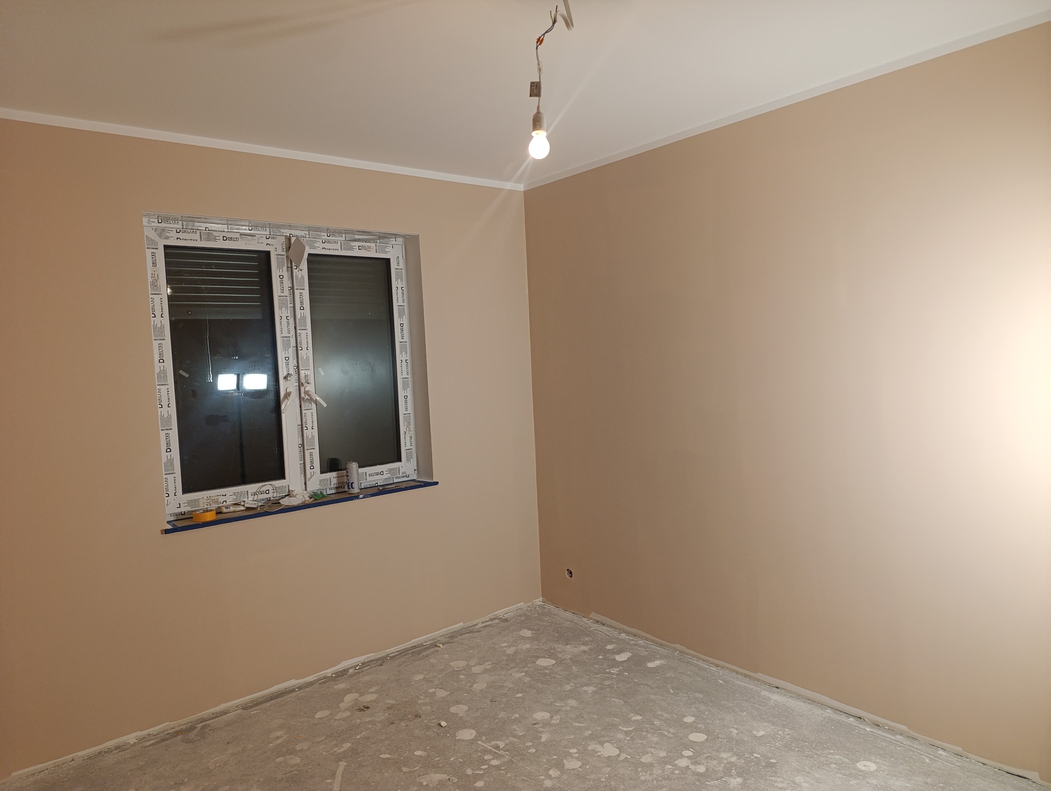 Pokój po gładziowaniu ścian na beżowo, widoczne okno z roletami, sufit z żarówką. Na parapecie narzędzia. Podłoga w plamach po pracach wykończeniowych.