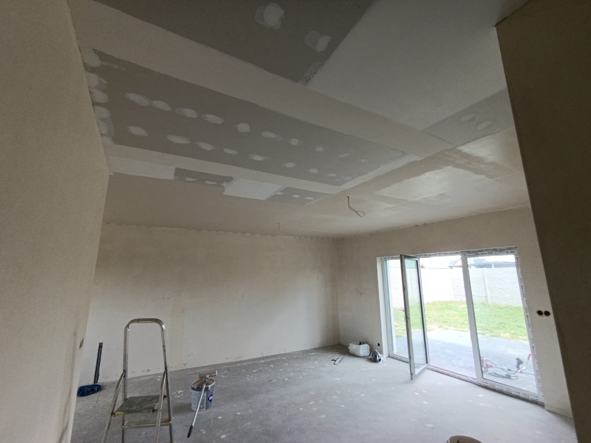 Wnętrze w trakcie prac wykończeniowych: ściany i sufit po szpachlowaniu, widoczna drabina malarska, otwarte okno balkonowe. Etap przygotowania do malowania.