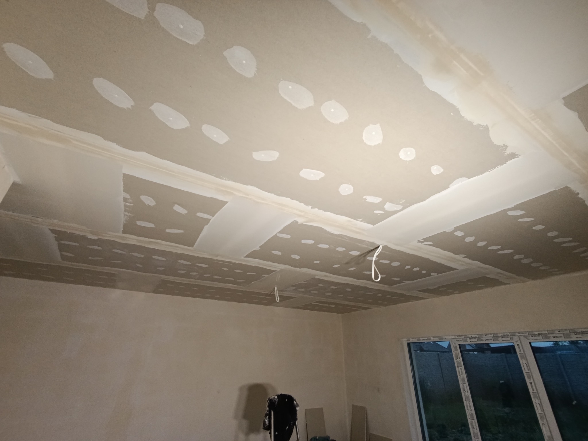 Sufit z płyt gipsowo-kartonowych w trakcie montażu, widoczne łączenia i otwory po wkrętach, przygotowane do szpachlowania. W tle okno i drabina.