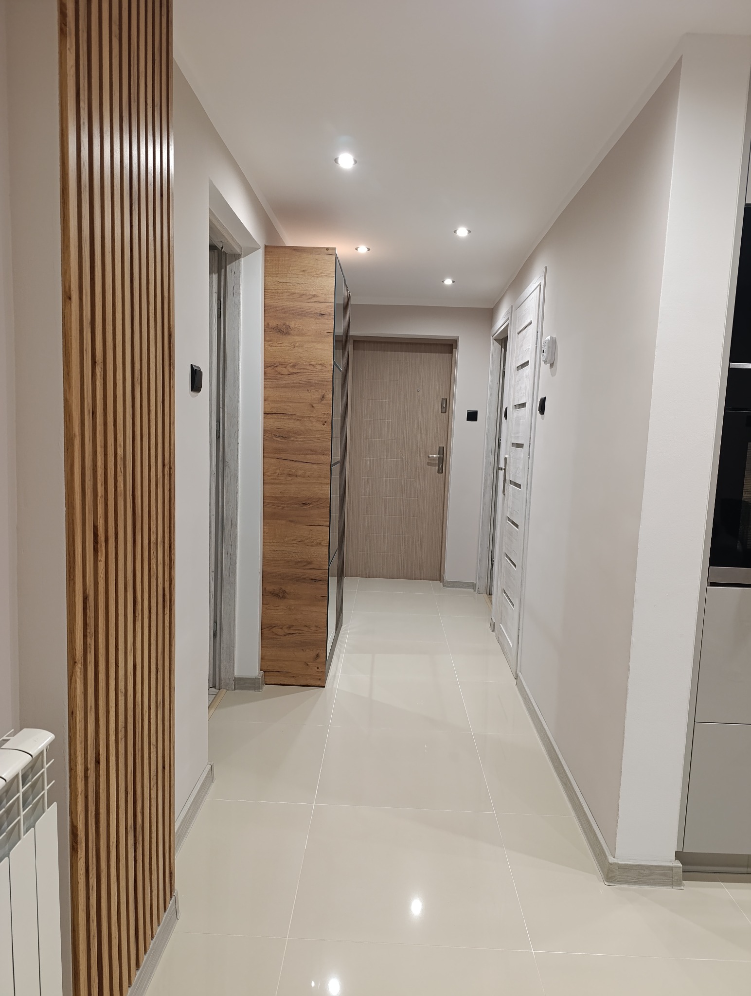 Nowoczesny korytarz po remoncie: drewniane lamele dekoracyjne, jasne płytki podłogowe, zabudowa meblowa z lustrem i drzwi. Oświetlenie punktowe.