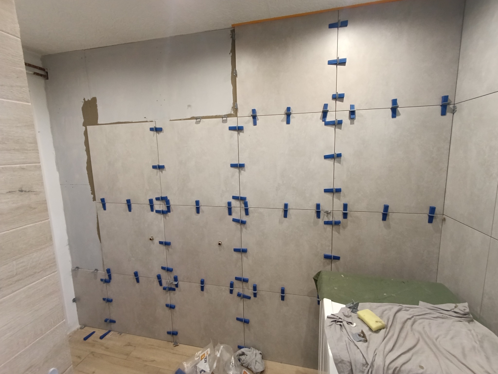 Ściana w trakcie układania płytek ceramicznych, z użyciem niebieskich klipsów poziomujących. Widoczne fragmenty niezakończonej powierzchni i narzędzia pracy.