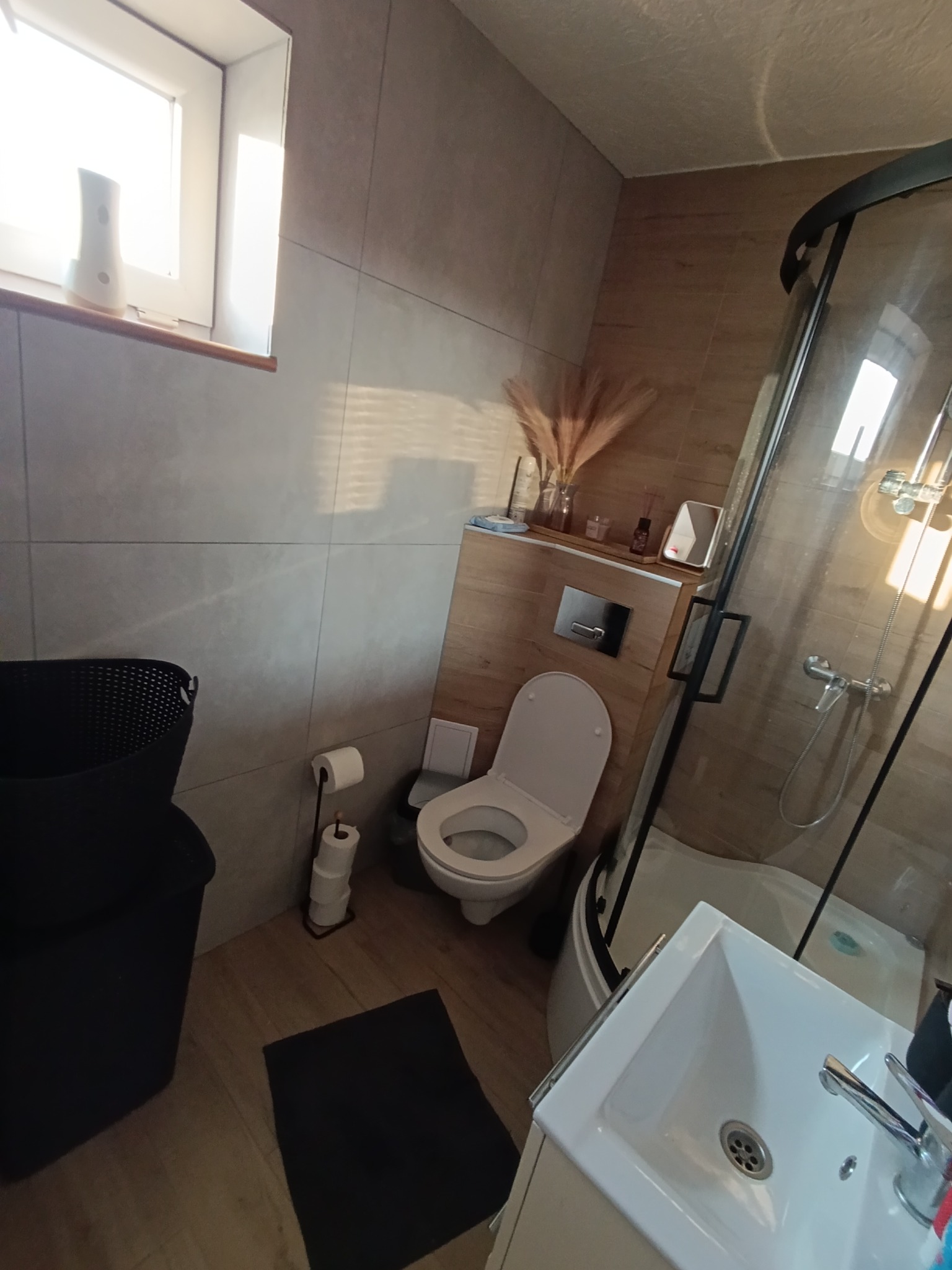 Jasna łazienka z kabiną prysznicową, toaletą i umywalką. Na ścianach płytki imitujące drewno i szare. Dekoracje na półce nad toaletą. Widok na okno z dyfuzorem zapachu.