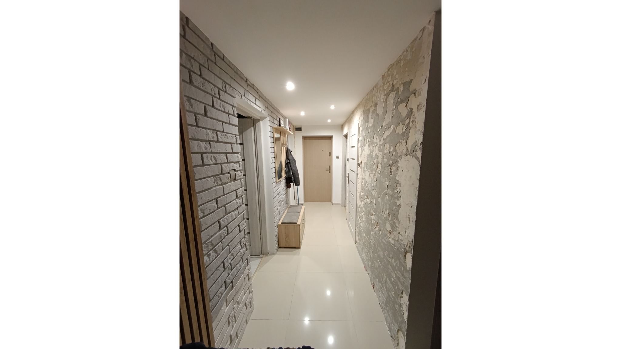 Korytarz w trakcie remontu: ściana z cegły dekoracyjnej po lewej, ściana w trakcie szpachlowania po prawej. Widoczne oświetlenie sufitowe i elementy wyposażenia.