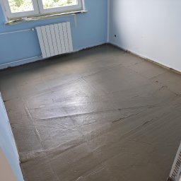 Perfect Home - Wylany, mokry podkład podłogowy w pokoju z niebieską ścianą i białym grzejnikiem. Widoczne ślady po rozprowadzaniu masy. Przygotowanie do dalszych prac wykończeniowych.