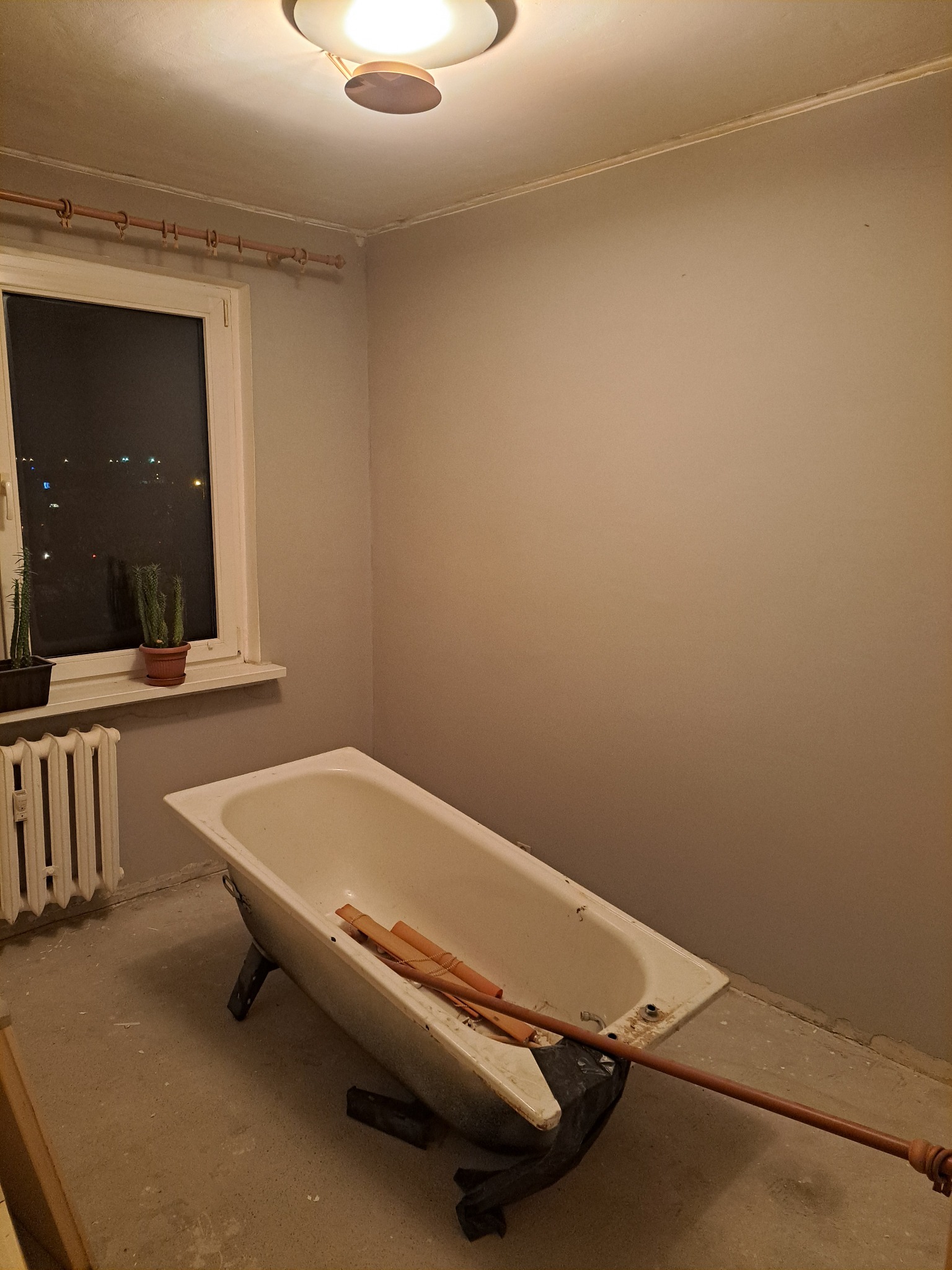Łazienka w trakcie remontu: stara wanna, grzejnik, okno z widokiem na nocne miasto. Ściany przygotowane do malowania, surowy beton na podłodze. Widoczne narzędzia.