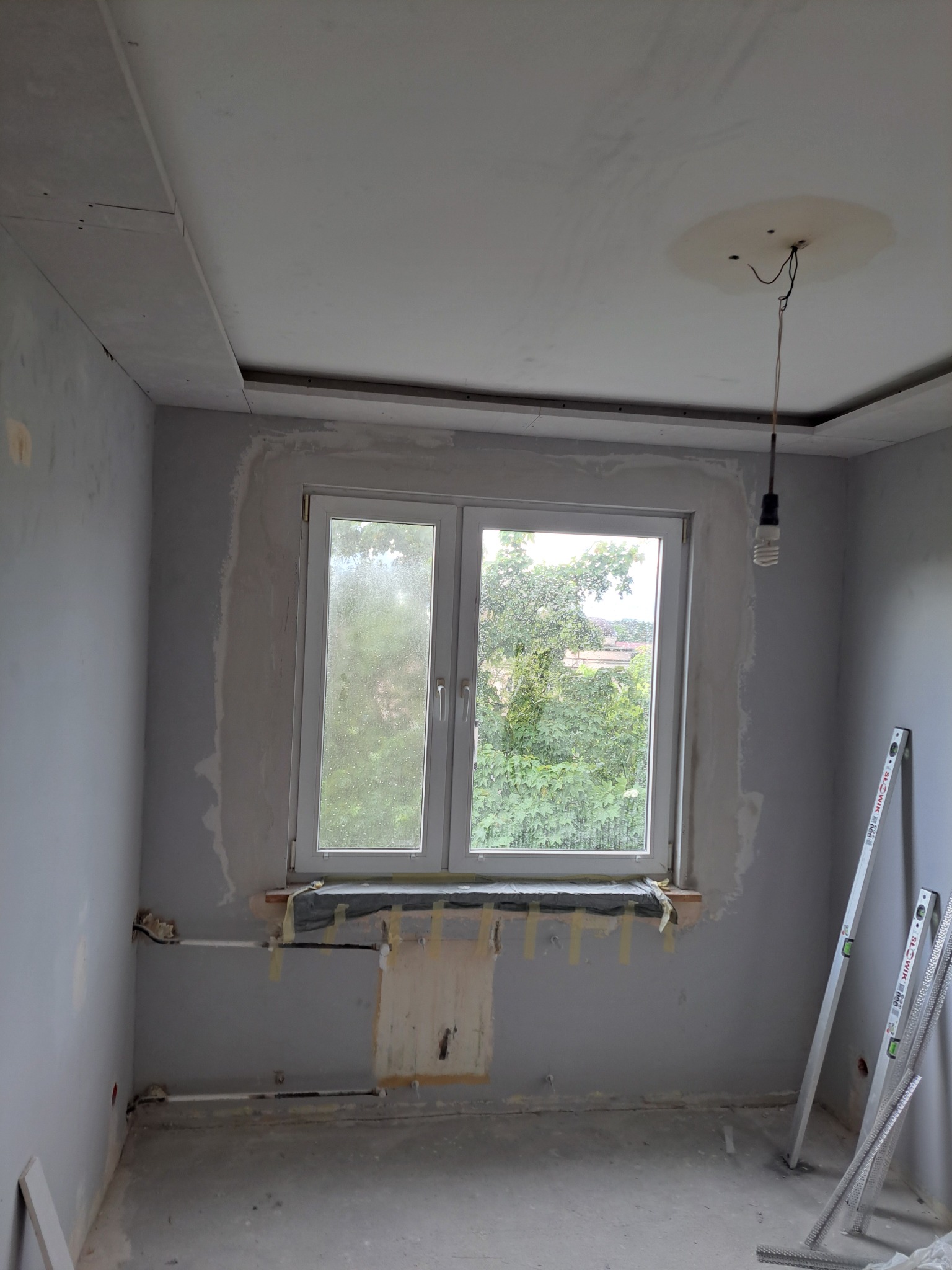 Pokój w trakcie prac wykończeniowych: szpachlowane ściany, sufit z płyt karton-gips, okno z widokiem na zieleń, poziomice oparta o ścianę. Widoczne ślady po instalacjach.