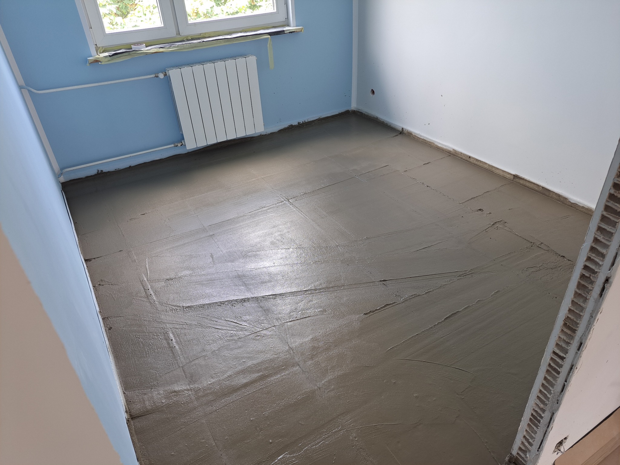Wylany, mokry podkład podłogowy w pokoju z niebieską ścianą i białym grzejnikiem. Widoczne ślady po rozprowadzaniu masy. Przygotowanie do dalszych prac wykończeniowych.