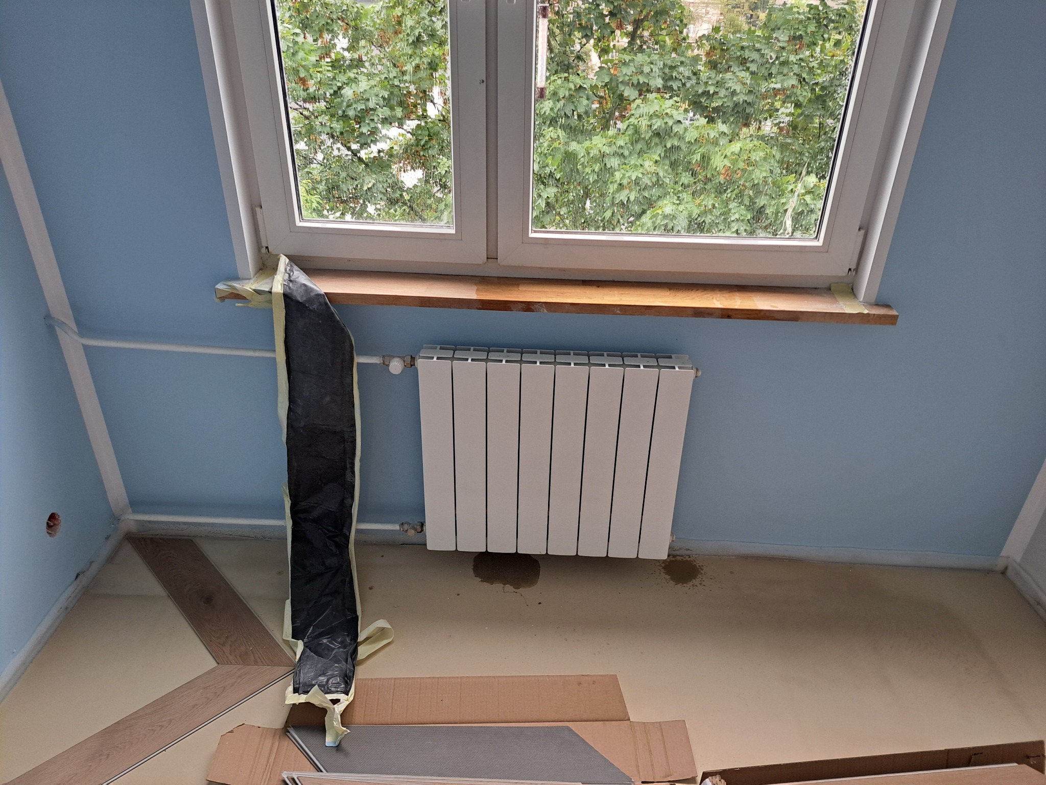 Pokój na poddaszu w trakcie prac wykończeniowych: okno dachowe, grzejnik, zabezpieczona folią rura, panele podłogowe w kartonach. Błękitne ściany i jasna podłoga.