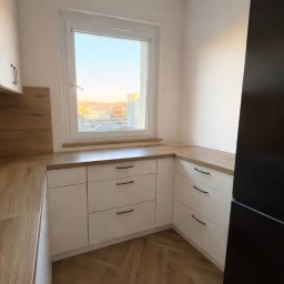 TIMBER CRAFT Krzysztof Windorpski - Jasna kuchnia na wymiar z drewnianym blatem i parkietem ułożonym w jodełkę. Widok z okna na miasto oświetlone zachodzącym słońcem. Minimalistyczne czarne uchwyty.