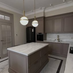 TIMBER CRAFT Krzysztof Windorpski - Elegancka kuchnia w odcieniach szarości z marmurowym blatem wyspy i podłogą. Dwie złote lampy wiszące nad wyspą, klasyczne fronty szafek i nowoczesne oświetlenie.