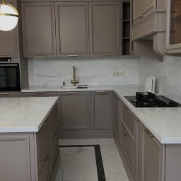 TIMBER CRAFT Krzysztof Windorpski - Elegancka kuchnia w odcieniach szarości z marmurowym blatem i złotymi uchwytami, oświetlona kulistą lampą. Widoczny fragment wyspy kuchennej i płyta grzewcza.