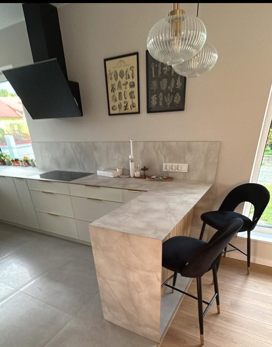 Nowoczesna kuchnia z wyspą wykończoną marmurkowym wzorem, czarne hokery, minimalistyczny okap i designerskie lampy wiszące. Jasne fronty szafek i blaty.