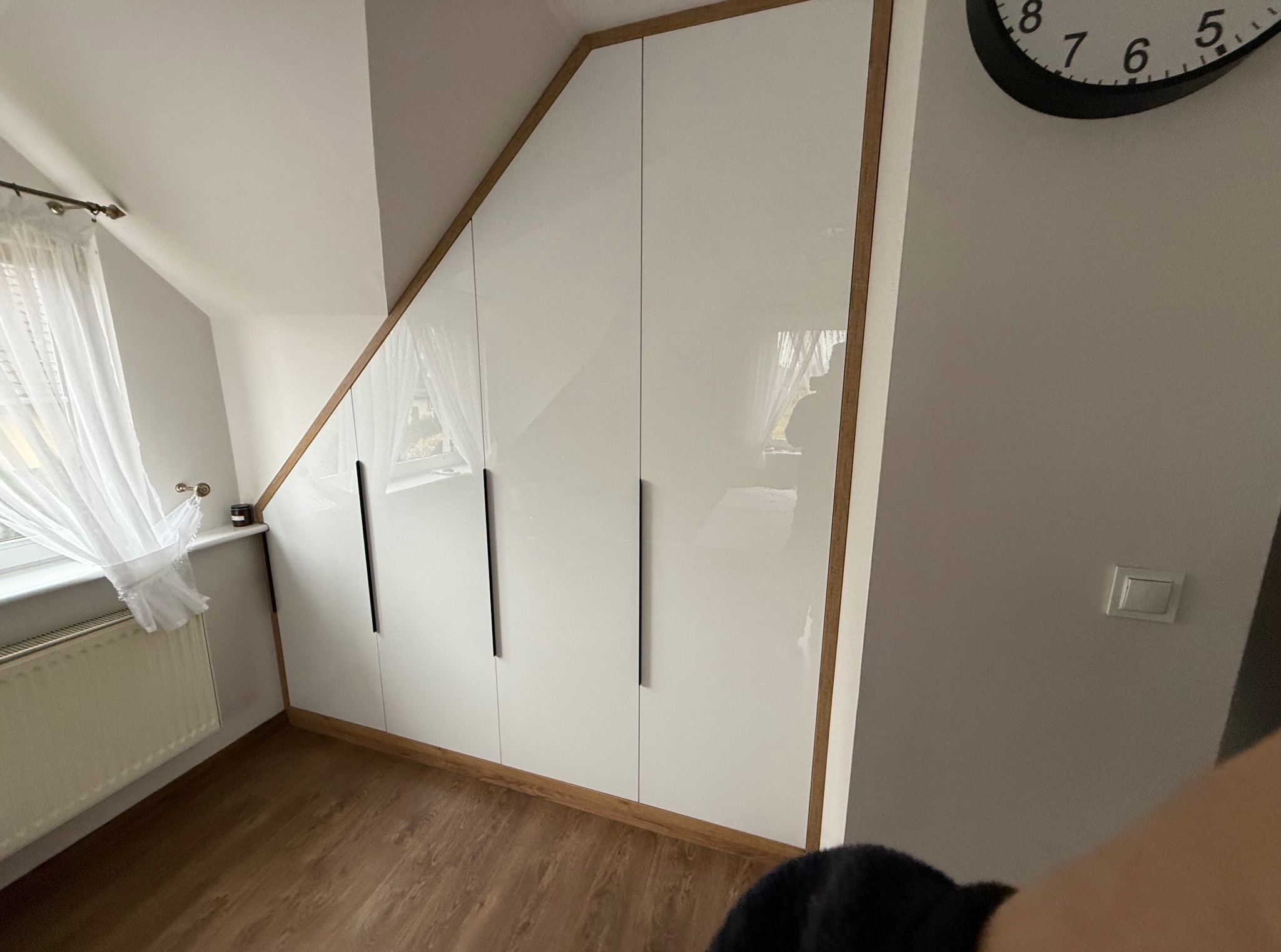 Biała szafa na wymiar z drewnianym obramowaniem, dopasowana do skosu poddasza. Widoczne czarne, podłużne uchwyty i fragment okna z firanką.